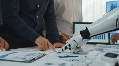 İş dünyasından profesyonellerden oluşan bir ekip, modern ofis ortamında veri tabloları ve raporlarını analiz ederek gelişmiş bir robot kol ile iletişim kuruyor.