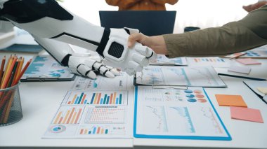 Çağdaş ofis ortamında bir robotla el sıkışan bir insan, analitik raporlar ve chartlarla çevriliyken teknoloji yoluyla işbirliği sergiliyor.