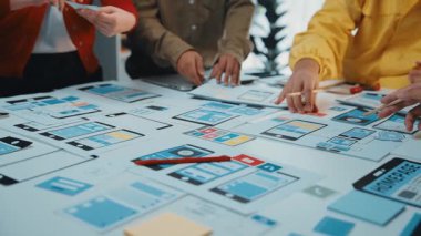 Dinamik bir ekip akıllı telefon arayüzü düzeni oluşturmak için beyin fırtınası yapıyor. Masaya yayılmış kağıtlar ve prototipler işbirliği ve yaratıcılığı teşvik ediyor. SACTR