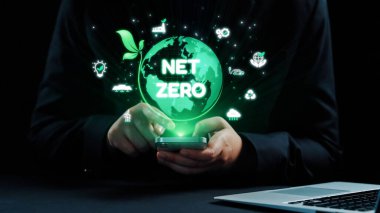 Bir kişi elinde net sıfır ve çevresel ikonlar içeren yeşil bir küreyi gösteren bir akıllı telefon tutuyor ve modern dünyada sürdürülebilirlik ve iklim sorumluluğuna bağlılığı simgeliyor. Dörtgen