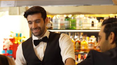 Güzel görünümlü barmen, içki tezgahındaki çiftle samimi bir gülümseme sohbeti yaparken aynı zamanda hoş geldin içkisi ve özel menü listesi öneren çekici profesyonellik. Vinosity.