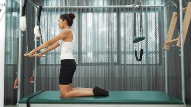 Kadın spor salonunda pilates reformatörüyle çekirdek güçlendirme egzersizi yapıyor. Çekici bayan pilates eğitmeni diz çökmüş vaziyette Cadillac 'ta omuz hareketliliğini arttırmak için bekliyor. Alışkanlık.