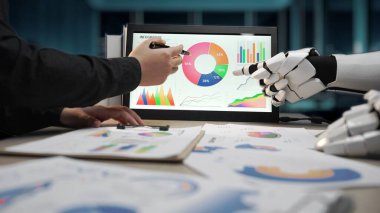 Bir iş adamı, modern bir ofis ortamında çeşitli grafik ve grafiklerin yer aldığı dijital ekranda sunulan verileri analiz ederek robotik bir asistanla işbirliği yapar. Noogenesis