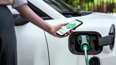 Akıllı telefon akü durumu arayüzünü elinde tutan iş kadını akıllı EV mobil uygulama ile otomobil parkındaki şarj istasyonundan elektrik şarj eden çevre dostu EV araba. İnceleyin