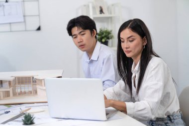 Profesyonel Asyalı erkek mimar ev modelinin uzunluğunu ölçmek için cetvel kullanırken, beyaz tenli genç meslektaşı ev modeliyle toplantı masasındaki verileri analiz etmek için dizüstü bilgisayar kullanıyor. Tertemiz..