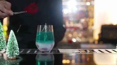 Lüks bar tezgahında kırmızı kurutulmuş meyve biberiyesiyle Noel Mavi Göl Kokteyli 'ni süsleyen yakın plan barmende yeşil neon kar ağacı şenlikli bokeh ışıklı neşeli bayram yer alıyor. Vinosity.