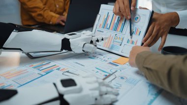 Detaylı iş raporları ve çizelgeleri üzerinde çalışan, ofis ortamında takım çalışmalarının ve teknolojinin geleceğini gösteren dinamik bir toplantı sahnesi. Noogenesis