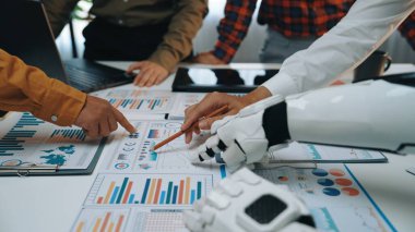İnsan elini ve robot kolunu gösteren modern bir ofis sahnesi performans raporlarını ve grafiklerini analiz eden, teknoloji ve insan görüşü arasındaki işbirliğini vurgulayan.
