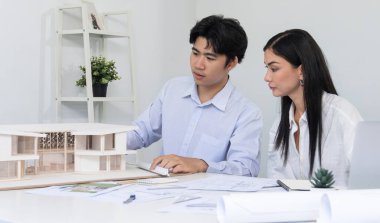 Profesyonel Asyalı erkek mimar ev modelinin uzunluğunu ölçmek için cetvel kullanırken, beyaz tenli genç meslektaşı ev modeliyle toplantı masasındaki verileri analiz etmek için dizüstü bilgisayar kullanıyor. Tertemiz..