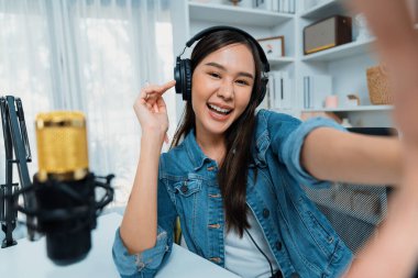 Sunucu kanalı genç ve güzel Asyalı kız akıllı telefonuyla selfie çekiyor. Modern stüdyo ofis konseptinde kulaklık takıyor. Sahne arkası yayıncısı sosyal medyada canlı yayın yapıyor. Taktik.