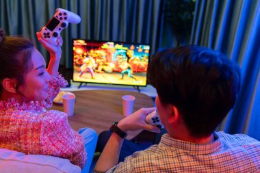 Joystickli birkaç oyuncu dövüş oyunu oynuyor, kadın oyuncu yarışması derecesi arka tarafta TV ekranında modern rahat evinde neon ışıklı oturma odasındaki koltukta oturuyor. İnfobahn.