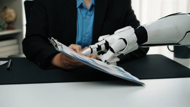 Bir robot el, modern ofis ortamında teknoloji ve insan etkileşiminin karışımını tasvir eden bir iş adamı tarafından tutulan bir panoyla etkileşir. Noogenesis