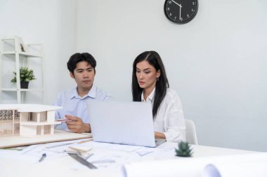Profesyonel Asyalı erkek mimar ev modelinin uzunluğunu ölçmek için cetvel kullanırken, beyaz tenli genç meslektaşı ev modeliyle toplantı masasındaki verileri analiz etmek için dizüstü bilgisayar kullanıyor. Tertemiz..