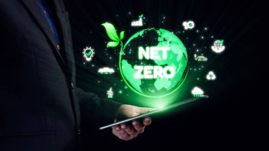 Takım elbiseli bir iş adamı, sürdürülebilirlik ve çevresel ilerlemeyi sembolize eden yeşil bir küre ve net sıfır metin içeren parlak bir tablet sunuyor. Dörtgen