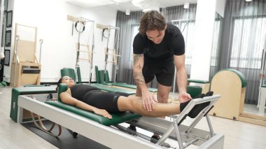 Mutlu pilates eğitmeni, bir müşteriye çekirdek istikrarı geliştirmek için bir reformcu üzerinde bacak egzersizi yapmasında yardımcı oluyor. Pilates makinesi kullanan sportif kız ark fıçısında kol dairesi pozu veriyor. Alışkanlık.