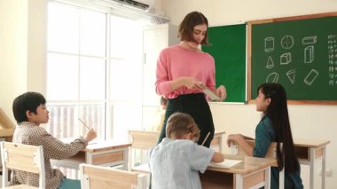 Zeki Asyalı öğrenci, ilkokulda öğretmen yürürken ve ödevlerini kontrol ederken matematik sorusunu sorarak elini kaldırdı. Öğretmen not defterini alırken soruyu cevaplıyor. Pedagoji.