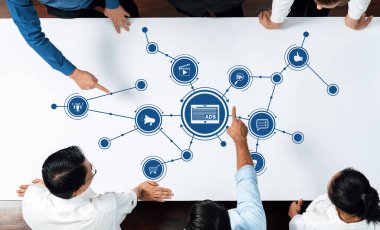 Çeşitli bir ekip, masadaki grafiksel reklam kavramlarına işaret ederek, pazarlama girişimlerinde takım çalışması ve stratejik planlama sergileyerek bir toplantıda işbirliği yapıyor. Dostluk