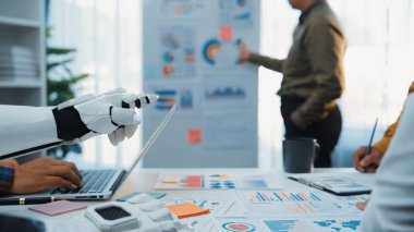 Gelecekçi bir robotik el dizüstü bilgisayar kullanırken bir işletme ekibi şık bir ofis ortamında grafikler ve grafikler üzerinde tartışıyor ve işbirliği sergiliyor.