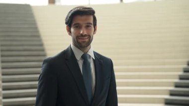 Akıllı iş adamı merdivenlerde kameraya bakıyor. İş elbisesi giyerken kameraya gülümseyen başarılı adamın yakın çekimi. Yönetici, kameraya bak. Hareket eden kamera. Sevinçli..