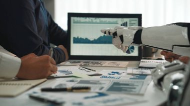 Profesyonel bir toplantı sırasında robot bir elin veri analiz çizelgelerine işaret ettiği modern bir çalışma alanı. Sahne, iş dünyasındaki yenilik ve işbirliğini simgeliyor. Noogenesis.