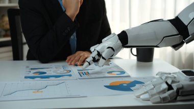 Robotik kol, bir iş toplantısı sırasında teknolojinin profesyonel analiz ve karar verme sürecine entegrasyonunu gösteren ayrıntılı raporlara ve çizelgelere işaret eder. Noogenesis