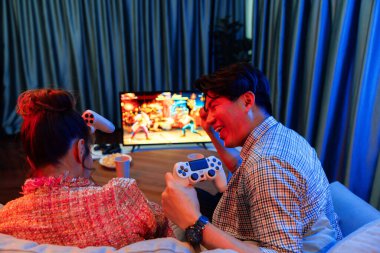 joystickli bir çift, TV ekranında dövüş oyunu oynuyor, erkek, kız arkadaşıyla yarışma seviyesini geri kazanıyor, modern evdeki neon ışıklı oturma odasında kanepeye oturuyor. İnfobahn.