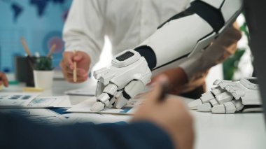 Robotik bir el ve insan eli, modern çalışma alanlarındaki otomasyon ve takım çalışmasını sembolize eden gösterişli bir ofis alanında proje planlamasında işbirliği yapmaktadır.