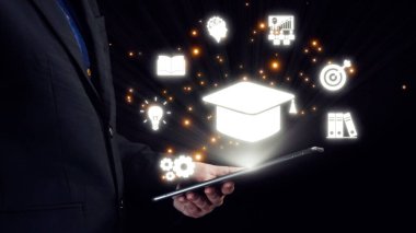 Bir işadamı, teknoloji ve öğrenmenin kesişimlerini gösteren çeşitli eğitim sembolleriyle aydınlatılmış bir tablet tutuyor. Görüntü yenilik ve bilgiyi temsil ediyor. Dörtgen