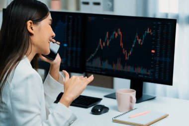 Genç ve güzel Asyalı iş kadını, modern ofisteki bilgisayar ekranlarında her borsa grafiğinde kârlı bir hisse senedi grafiğiyle istikrarlı veya daha yüksek kar değerini analiz etmek için komisyoncuyu telefonla arıyor. Taktik.