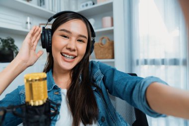 Sunucu kanalı genç ve güzel Asyalı kız akıllı telefonuyla selfie çekiyor. Modern stüdyo ofis konseptinde kulaklık takıyor. Sahne arkası yayıncısı sosyal medyada canlı yayın yapıyor. Taktik.