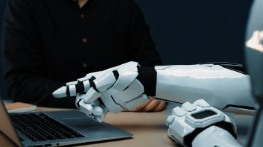 Günümüz dijital dünyasında insanlar ve gelişmiş teknolojiler arasındaki işbirliğini gösteren, modern çalışma alanındaki bir dizüstü bilgisayarı gösteren bir robot elin yakın çekimi. Noogenesis