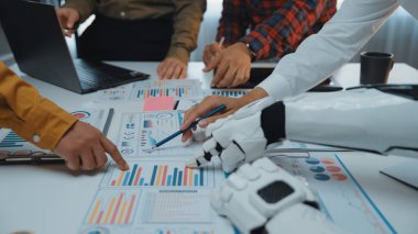 İnsan eliyle robot kol arasındaki işbirliğini anlatan modern bir ofis sahnesi. İş haritaları ve grafikleri incelenirken teknoloji ve takım çalışmasının karışımını gösteriyorlar. Noogenesis