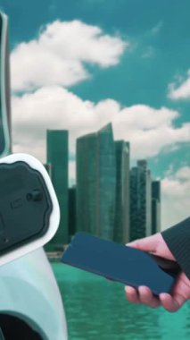 İlerici işadamı elektrikli aracı şarj kablosu ile şarj etme istasyonu ve temiz enerji kampanyası için şehir arka planına sahip akıllı telefondan enerji durumunu kontrol etme.