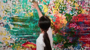 Genç ve güzel bir kızın arka görünüşü renkli duvarı boyamak için elini kaldırır. Çekici bir lise öğrencisi renk paletini tutarken duvarı el iziyle boyuyor. Yaratıcı aktivite. Öğretim