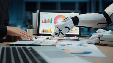 Bir robot el, modern bir ofisteki çizelge ve grafikleri kullanarak iş verilerini analiz etmek için bir insanla işbirliği yapar ve teknolojinin takım çalışmasındaki rolünü vurgular. Noogenesis