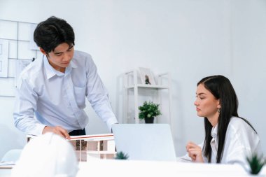 Profesyonel Asyalı erkek mimar ev modelinin uzunluğunu ölçmek için cetvel kullanırken, beyaz tenli genç meslektaşı ev modeliyle toplantı masasındaki verileri analiz etmek için dizüstü bilgisayar kullanıyor. Tertemiz..