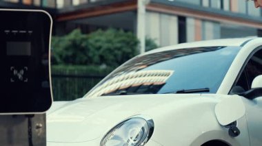 İlerici işadamı, şehir merkezinde dolaşmadan önce şarj cihazının fişini şarj istasyonundan elektrikli arabasına çekiyor. Çevre dostu şarj edilebilir araba, sürdürülebilir ve temiz enerjiyle çalışıyor..