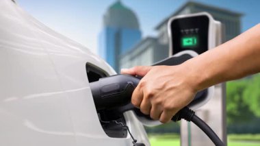 Adam elektrikli arabaya akıllı EV şarj cihazı takıyor ve arka planda modern şehir manzarası olan EV şarj istasyonundan şarj oluyor. ESG şehrinde gelecekteki sürdürülebilir güç ve EV araba kullanımı. İnceleyin