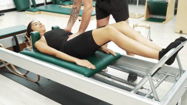 Mutlu pilates eğitmeni, bir müşteriye çekirdek istikrarı geliştirmek için bir reformcu üzerinde bacak egzersizi yapmasında yardımcı oluyor. Pilates makinesi kullanan sportif kız ark fıçısında kol dairesi pozu veriyor. Alışkanlık.