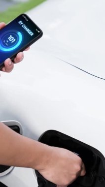 Modern yaşam tarzı adamı EV şarj cihazını ev şarj istasyonuna yerleştirir ve akıllı telefondan EV batarya durumunu etkinleştirir. Gelecekçi enerji sürdürülebilirliği ve elektrikli araba. İnceleyin