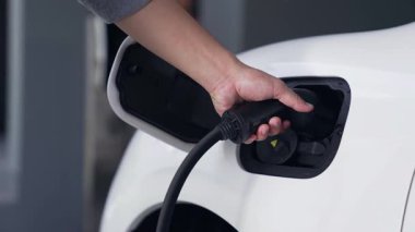 Adam elektrikli arabasını akıllı ev şarj istasyonundaki EV şarj cihazıyla şarj ediyor. Fütürist dijital batarya durumu hologramı. Sürdürülebilir güç için EV araba ve ev enerjisi altyapısı. İnceleyin