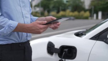Takım elbiseli, hırslı bir işadamı elektrik kablosu ve yenilenebilir enerji enerjili bir elektrikli araçla şarj istasyonunda dururken elektrikli aracına yaslanıyor..