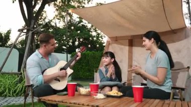 Baba gitar çalarken anne ve kız birlikte kampta oturuyorlar. Ebeveynler, genç nesil çapraz kuşak farklılıklarıyla zaman geçirmek için açık hava kamp aktivitelerini kullanıyorlar. Uyumsuzluk.