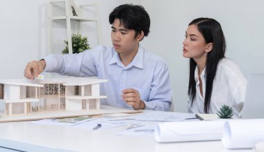 Profesyonel Asyalı erkek mimar ev modelinin uzunluğunu ölçmek için cetvel kullanırken, beyaz tenli genç meslektaşı ev modeliyle toplantı masasındaki verileri analiz etmek için dizüstü bilgisayar kullanıyor. Tertemiz..