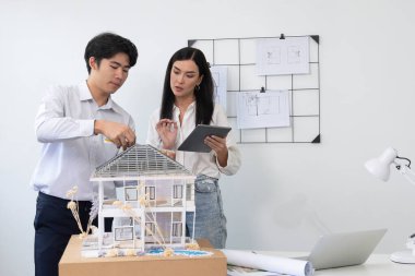Güzel, genç, beyaz bir proje yöneticisi Asyalı mühendise, mühendislik masasında ev modelini ölçerken tablet kullanarak bina detaylarını anlattı. Tertemiz.