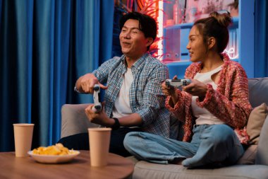 Neşeli, gülümseyen bir çiftin koltuğunda oturup oyun oynuyorlar. Oyun konsolu, hazır aperatifler ve içecekler, modern neon ışıklı, rahat oturma odasında eğlenceli bir rekabet seviyesinde. İnfobahn.