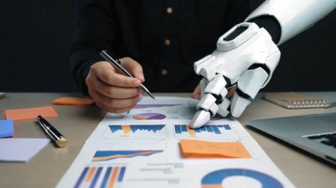 Bir insan eli ve robot el, modern bir çalışma alanında teknoloji ve insan zekası arasındaki sinerjiyi gösteren veri analizi üzerinde birlikte çalışırlar. Noogenesis