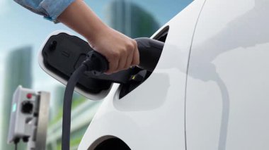 Adam elektrikli arabaya akıllı EV şarj cihazı takıyor ve arka planda modern şehir manzarası olan EV şarj istasyonundan şarj oluyor. ESG şehrinde gelecekteki sürdürülebilir güç ve EV araba kullanımı. İnceleyin