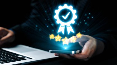 Bir iş adamı, yıldızlı bir dijital ödül ikonunu gösteren, modern ofis ortamındaki başarı ve kaliteyi sembolize eden bir akıllı telefonla bağlantı kuruyor. Dörtgen