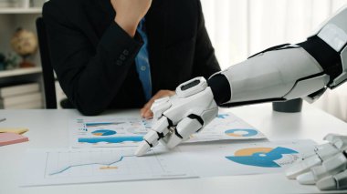 Bir robot kol, ofis masasındaki detaylı veri tablolarını ve grafikleri incelemek için bir işadamıyla işbirliği yapıyor. İş ortamlarında gelecekteki teknolojileri gösteriyor. Noogenesis